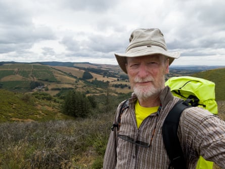 Selfie of a bearded man in a hat with bright yellow backpack in front of a landscape of low hills