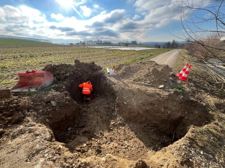 A person in a neon pink work jacket stands inside a huge hole next to mounds of dirt with a road in the background.