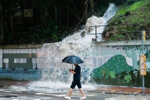 A man with an umbrella walks past a torrent of water cascading down a hillside and over a wall into the street