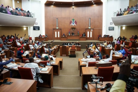 People in a debating chamber raising their hands to vote or speak. They sit at desks set in a semicircle facing two rows of officials, watched by people in an upper viewing gallery