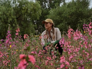 A woman wearing braids, a ball cap, and gloves holds a trowel and sits among wildflowers