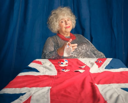 Portrait of Miriam Margolyes holding a broken union flag teacup while sitting at a table with a union flag tablecloth