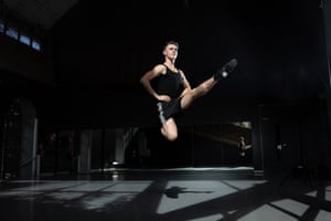 Liam Costello is photographed in mic-air as he dances in a dark studio. He has his hands on his hips, with one knee bent and the other leg doing a high kick. His body is illuminated in light but the mirrored studio is dark