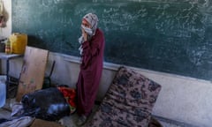 A woman standing in front of a blackboard pulls her headscarf over her mouth as she cries.