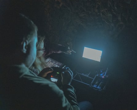 In a dark room a man holds a control panel while a woman points at a small screen