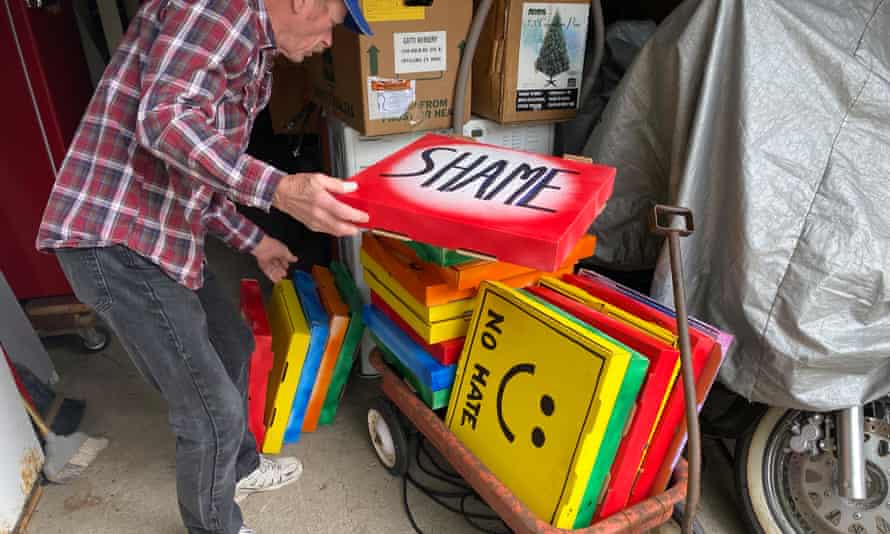 A man in a red plaid shirt sifts through brightly painted pizza boxes stacked on an old red metal wagon. One reads 'Shame' and another in yellow has a smiley face with 'No hate' written below.