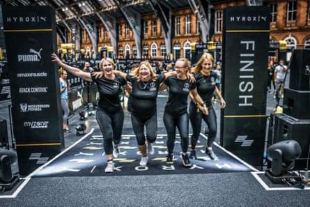 Four women in black T-shirts and leggings cross the finish line at a Hyrox event, arms over each others’ shoulders
