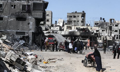 Palestinians push carts or wheelchairs of belongings past rubble and destroyed buildings in Khan Younis, southern Gaza, against a backdrop of a blue sky. 