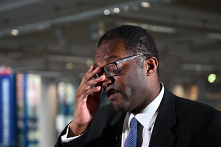 Kwasi Kwarteng, wearing a suit and tie, holds his hand to his face during a television interview