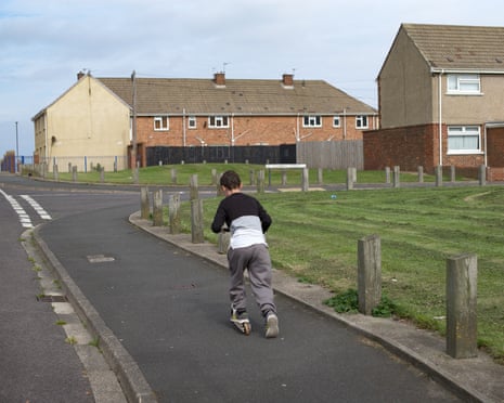 A child scooters along a pavement past wooden posts and grass, with residential houses in the background