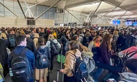 Passport control at Stansted airport: a large and disorganised-looking crowd of people are crammed into an airport building; they are carrying rucksacks and holding wheelie cases, and some are holding their phones. They are seen from behind and there are no discernible queues or lines.
