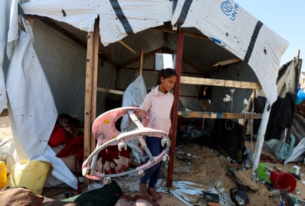 A girl in a pink sweatshirt stands outside a damaged makeshift tent, holding a baby walker. Possessions are strewn around in the sand.