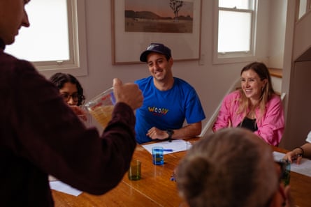 A man and woman sit at a dining table, smiling, while their friend pours them a glass of ginger beer.