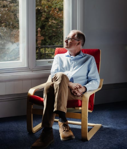 A man in a blue shirt looks out of a window while sitting in a red-upholstered armchair.