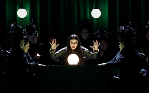 A performer, with long dark hair and makeup, sits behind a glowing white orb with her hands raised. Other performers, women in black costume with feathered headwear, sit around her