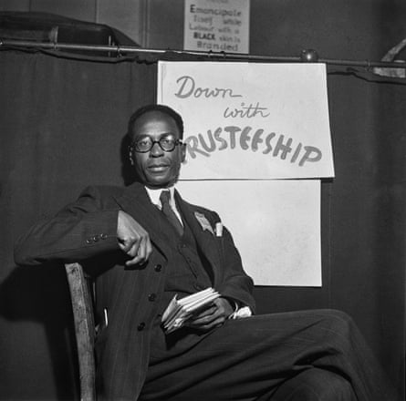 Black-and-white photo of a suited man sitting in front of a poster that reads ‘Down with trusteeship’
