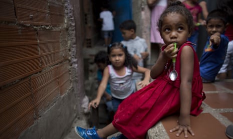 A young girl sits on a wall in a slum neighbourhood with a spoon in her mouth