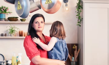 A woman holding a young girl and standing in a kitchen with yellow toxic stickers on fruit in the bowl, bottles of cleaning products and saucepans hanging from the ceiling