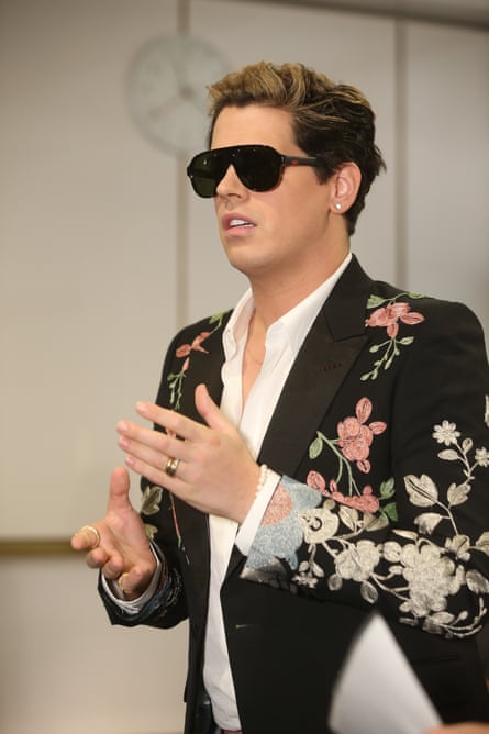 A young man with frosted brown hair, black fashionable sunglasses, a white collared shirt open at the neck, and a jacket decorated with white and pink flowers.