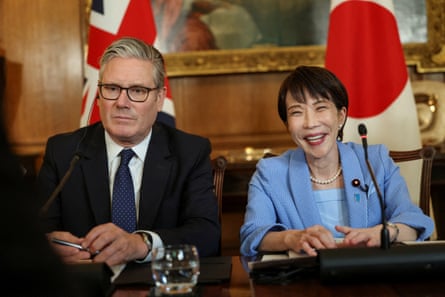 Keir Starmer and Sanae Takaichi sit together behind a large wooden table with the UK and Japan flags behind them. Starmer looks stern but Takaichi, who is wearing a pale blue jacket and top, is smiling widely.