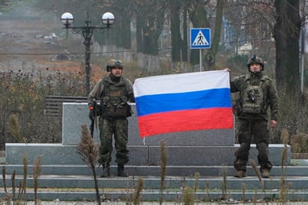 Russian soldiers hold a Russian national flag: they are standing on steps at what appears to be a stone monument or memorial in a park on a grey, wet, wintry day. The white, blue and red striped flag is bright in the gloom.