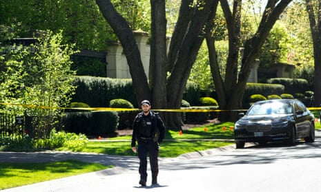 a man in a black uniform walks on a sunny street lined with green manicured lawns and tall trees and a walled off property