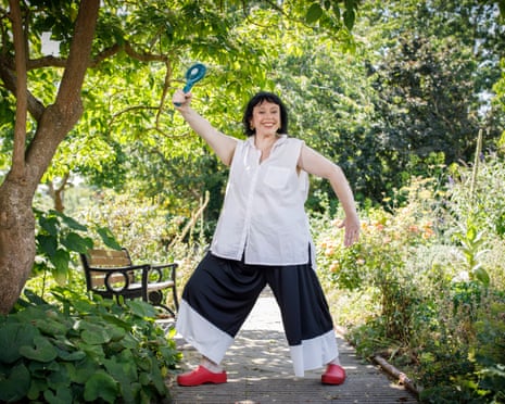 Amy Annette in a white shirt and wide black trousers strikes a playful pose in a garden, holding a blue hat aloft