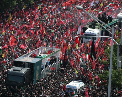 A truck holding the coffins of the former supreme leader and family members moves through a crowd of people holding flags.