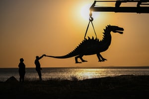 Silhouette image showing a large metal dinosaur sculpture being hoisted by a crane at sunset in Van, Turkey.