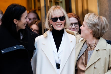 An older blond woman in sunglasses smiles as other women embrace her.