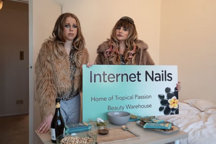 Nikki and Donna, in fur coats and wigs, hold an Internet Nails sign at a table