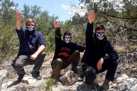 Three young white men, all with the lower halves of their faces covered by masks that make them look like skeletons, squatting on rocks in a forest and raising their right arms in the Nazi salute.