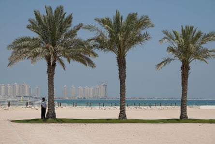 A security guard on a beach stands in the shade of a palm tree.
