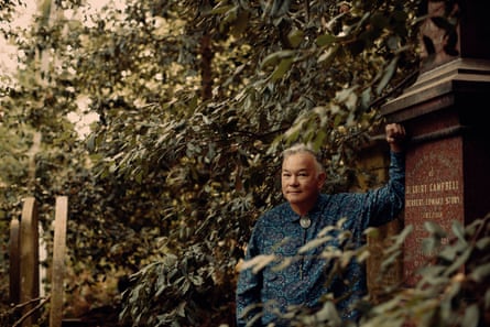Stewart Lee standing in a leafy cemetery with he left arm bent and his left hand leaning high up on a gravestone