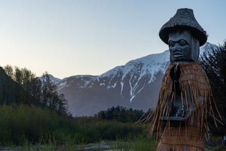 A huge pole with a face carved into it and wearing a hat, cloak and skirt woven from straw stands in the foreground. Behind is the river and a snow-capped mountain