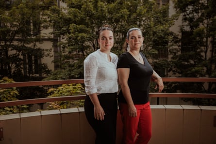 Two women with dark hair pulled back in pony tails, standing on a balcony with trees behind them