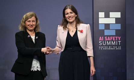 Věra Jourová and Michelle Donelan shake hands and smile next to a sign reading 'AI safety summit'. Both are smartly dressed, Jourová wearing a black jacket over white top with lace-like detailing and Donelan a pale pink jacket with black top and loose-fitting trousers