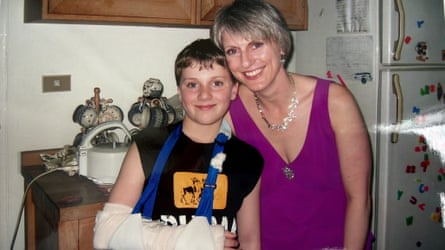 A boyin a black T-shirt with his arm in a plaster cast. His mum is standing by him, wearing a fuschia-coloured top. They are standing in a kitchen.