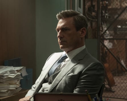 John Hamm sitting on a chair in a room, surrounded by papers, with a metal gate behind him
