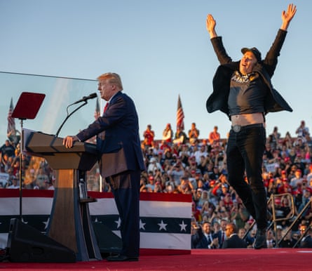 Elon Musk, in a baseball cap, jumps in the air with arms raised, his T-shirt riding up over his jeans, behind Donald Trump onstage at a rally