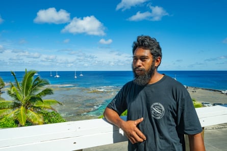 A bearded man stands in front of a clear blue sea and palm tree.