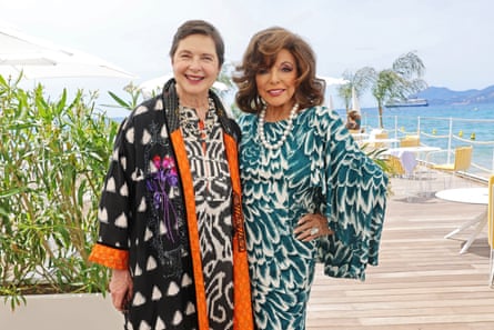 Isabella Rossellini and Joan Collins in Cannes. They are standing on wooden decking at the waterfront and both wear extravagantly patterned kaftan-style dresses with graphic black and white and green and white prints. Rossellini also wears a black kimono with white triangle pattern and orange edging.