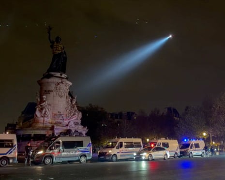 Police vans line Paris's Place de la République at night during curfew, a searchlight beam crossing the sky
