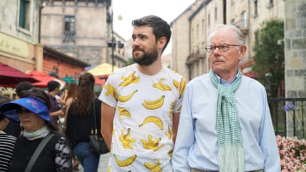 Walking in a street, him in a white T-shirt with bananas pictured on it; his father in a blue shirt and scarf