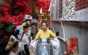 A woman in a floral headdress rides a moped down a narrow street past other women in similar attire