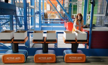 A queue of flat-topped robots carrying boxes wait while a female worker fills the boxes up