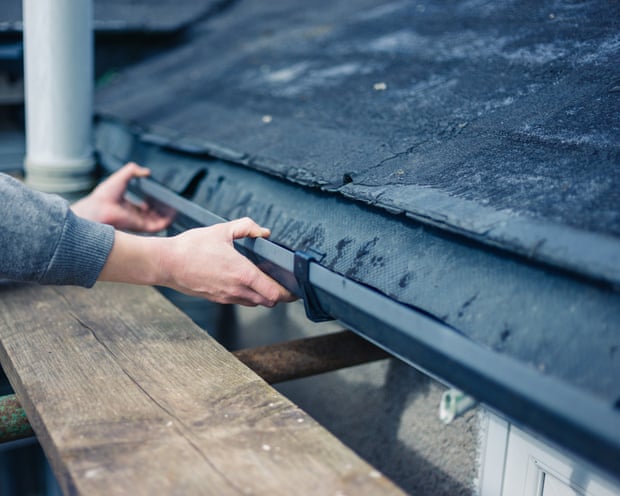 A worker's hands fit a guard onto a gutter beneath a slate roof from scaffolding