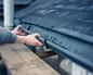 A worker's hands fit a guard onto a gutter beneath a slate roof from scaffolding