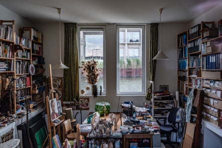 A room with bookcases filled with books on one wall and files and folders on the opposite wall. there is a workstation table with paintings on in the middle of the room and two long vertical windows overlooking the water