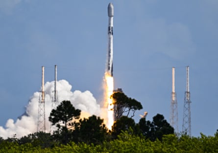 A rocket takes off into a clear blue sky with flames and clouds of white smoke shooting from beneath it